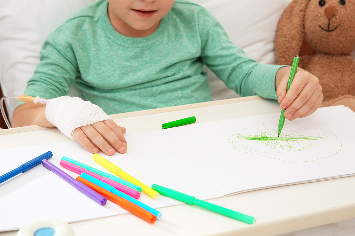 Kleines Kind mit Infusion am Arm malt ein Bild in einem Krankenhausbett